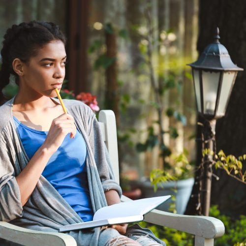 Cheerful charming african young girl dressed in a blue t-shirt and grey jacket holds the pencil at her hand and writing at the notebook on the nature background.